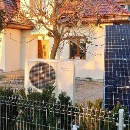 Instalacje Tomasz Magnuszewski - Biała pompa ciepła Fujitsu i panele fotowoltaiczne na tle domu jednorodzinnego, widoczne przez metalowe ogrodzenie. Słoneczny dzień.