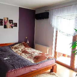 Sypialnia z czarną klimatyzacją Haier Serene Black nad łóżkiem. Ściana w pasy fioletu i szarości. Okno balkonowe z firanką. Roślina doniczkowa obok.