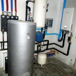 Instalacja grzewcza w kotłowni: srebrny zasobnik ciepłej wody Galmet z termometrem, biały bojler, niebieski zbiornik wyrównawczy, piec Kaisai, czarne rury i zawory, czerwone i niebieskie rurki...