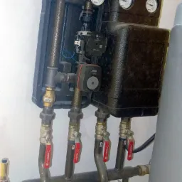 Czarna skrzynka rozdzielcza z manometrami, pompą obiegową i zaworami odcinającymi na rurach instalacji grzewczej, widoczne marki Watts, Grundfos i Ferro.