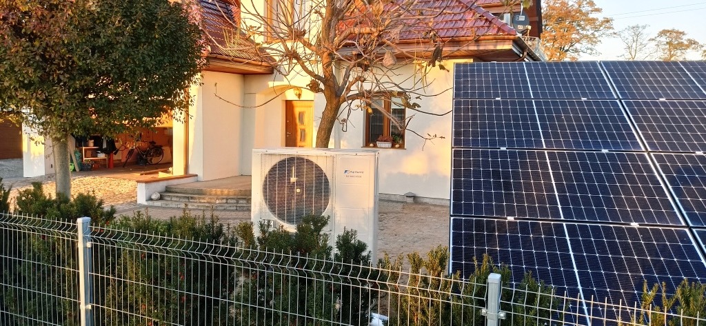 Biała pompa ciepła Fujitsu i panele fotowoltaiczne na tle domu jednorodzinnego, widoczne przez metalowe ogrodzenie. Słoneczny dzień.