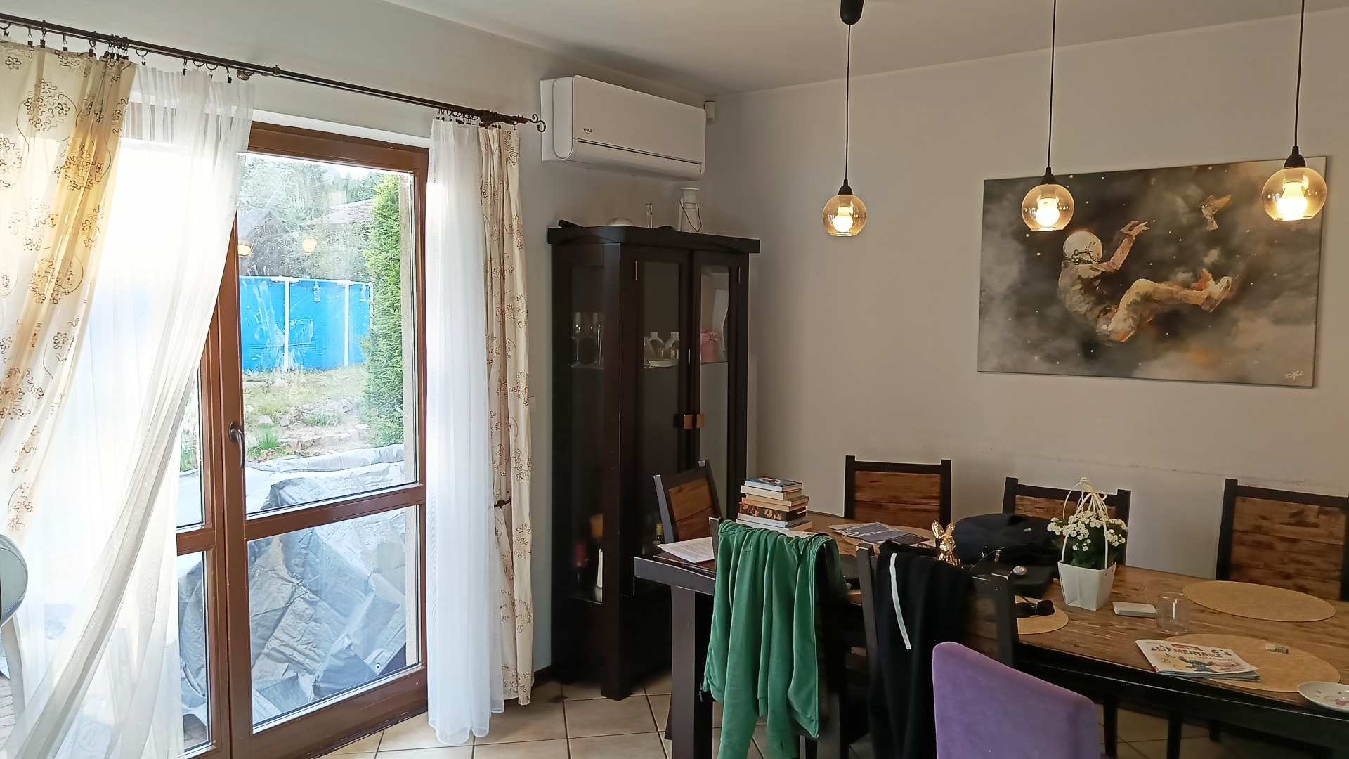 Wnętrze pokoju z klimatyzatorem Vivax H+ zamontowanym nad ciemnym kredensem. Widoczne okno z firankami i obraz na ścianie.