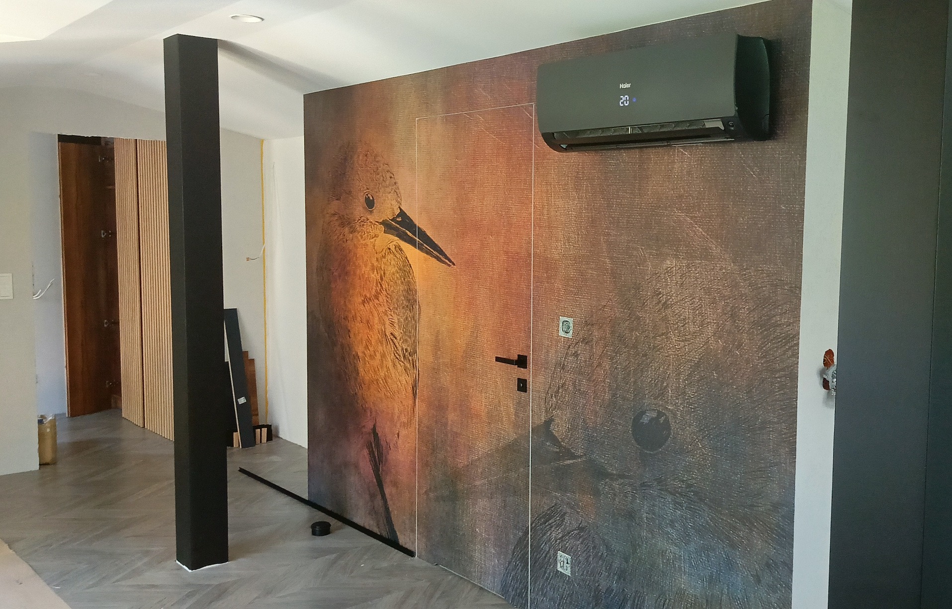 Czarna klimatyzacja Haier Flexis na ścianie z motywem ptaka, w nowoczesnym wnętrzu. Widoczny fragment kolumny i drzwi.