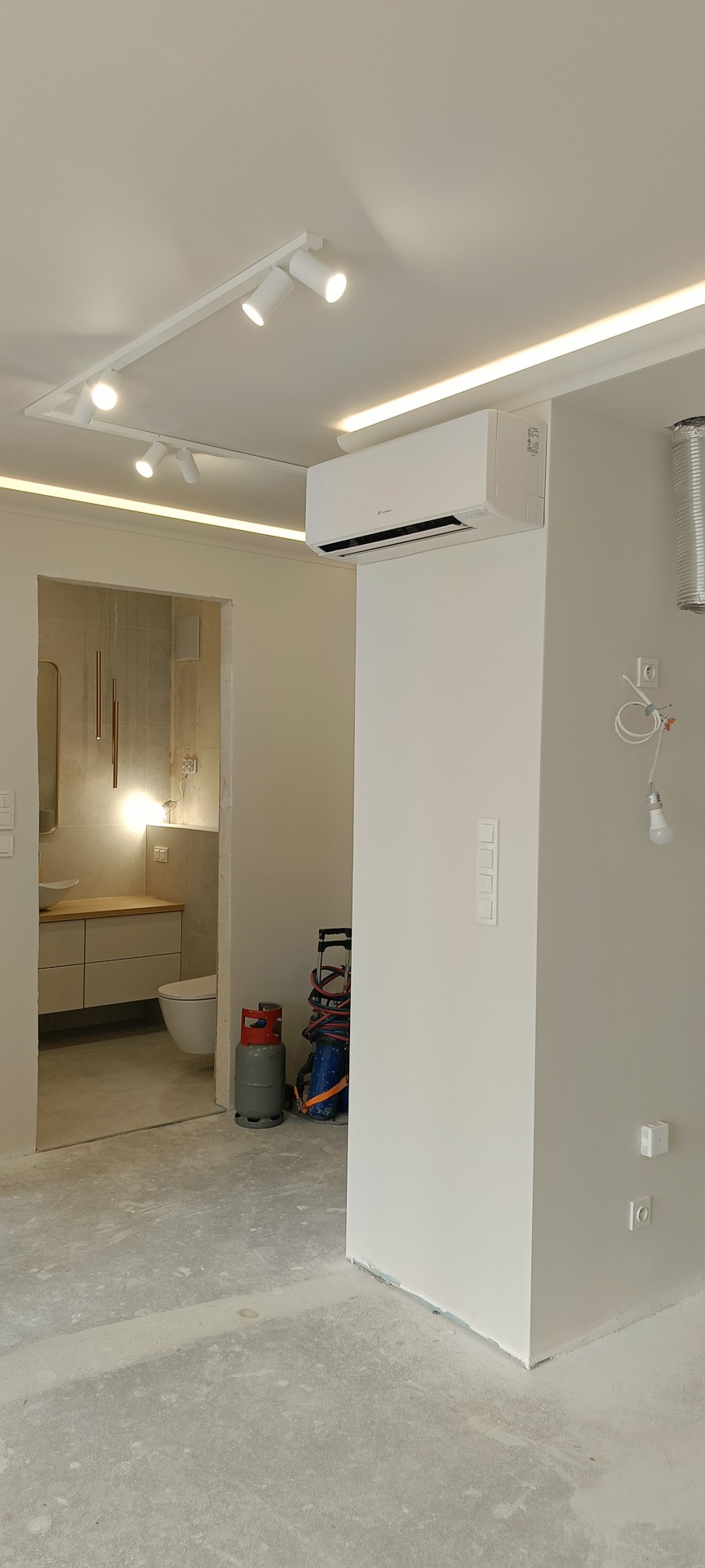 Biały klimatyzator Fuji KN zamontowany na ścianie w jasnym wnętrzu. Widoczne oświetlenie sufitowe i fragment łazienki w tle. Na podłodze butla gazowa i narzędzia.
