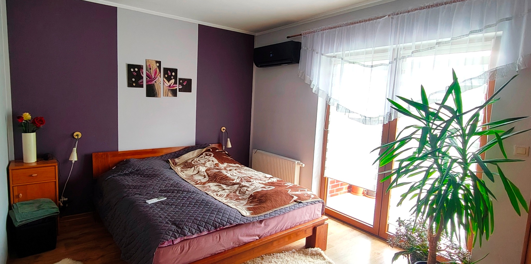 Sypialnia z czarną klimatyzacją Haier Serene Black nad łóżkiem. Ściana w pasy fioletu i szarości. Okno balkonowe z firanką. Roślina doniczkowa obok.