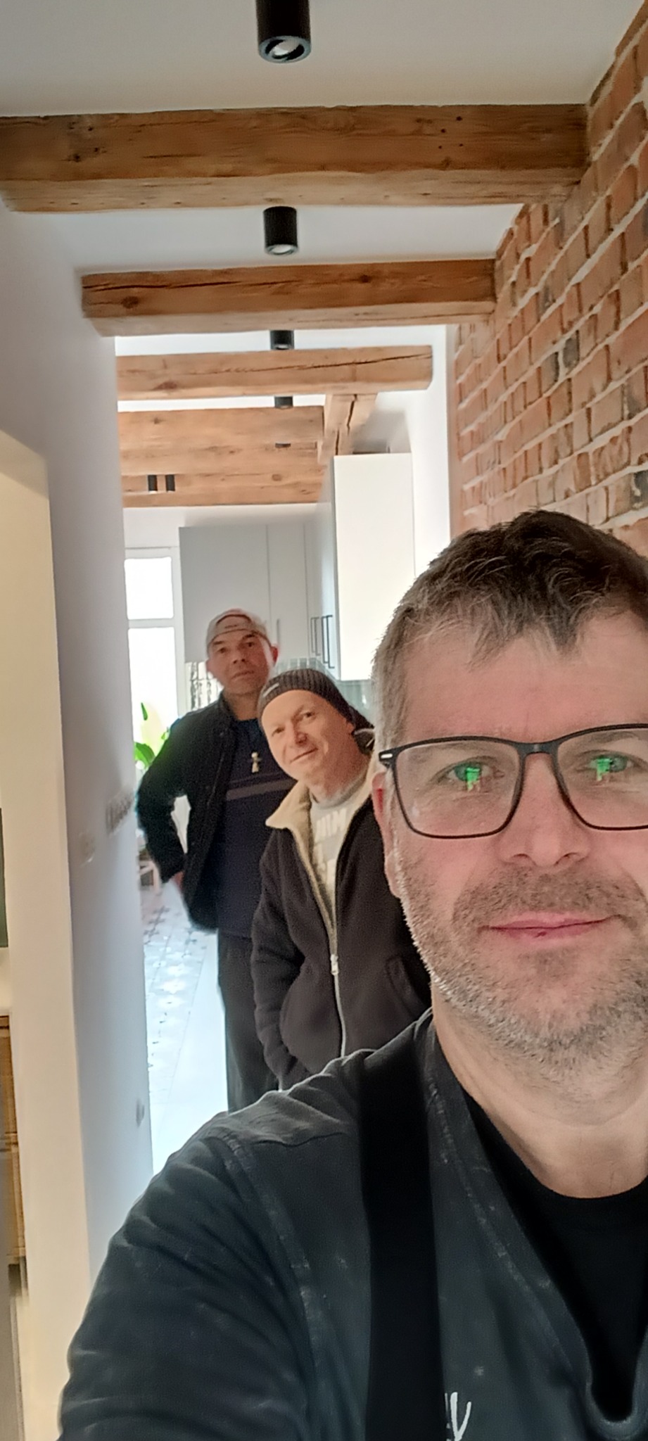 Trzech mężczyzn w korytarzu z drewnianymi belkami i ceglaną ścianą. Widoczne oświetlenie punktowe. Selfie jednego z nich.