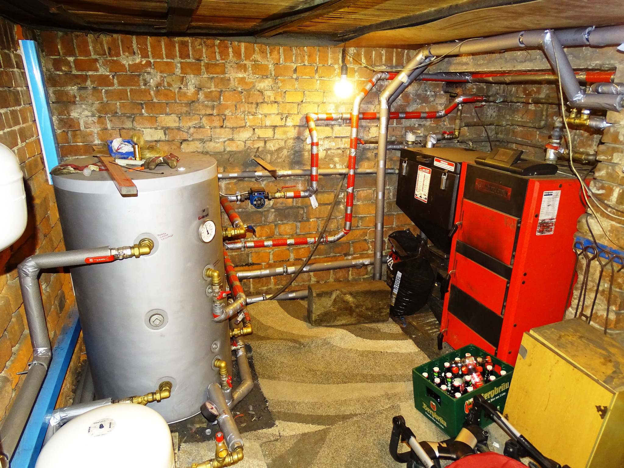 Piwnica z kotłem grzewczym, bojlerem i instalacją hydrauliczną z rurami w izolacji, dodatkowo skrzynka piwa i wózek dziecięcy.