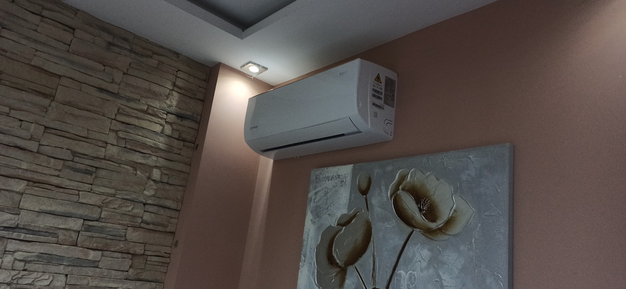 Biały klimatyzator ścienny zamontowany w rogu pokoju, obok ściany wyłożonej kamieniem dekoracyjnym i obrazu z motywem kwiatowym.