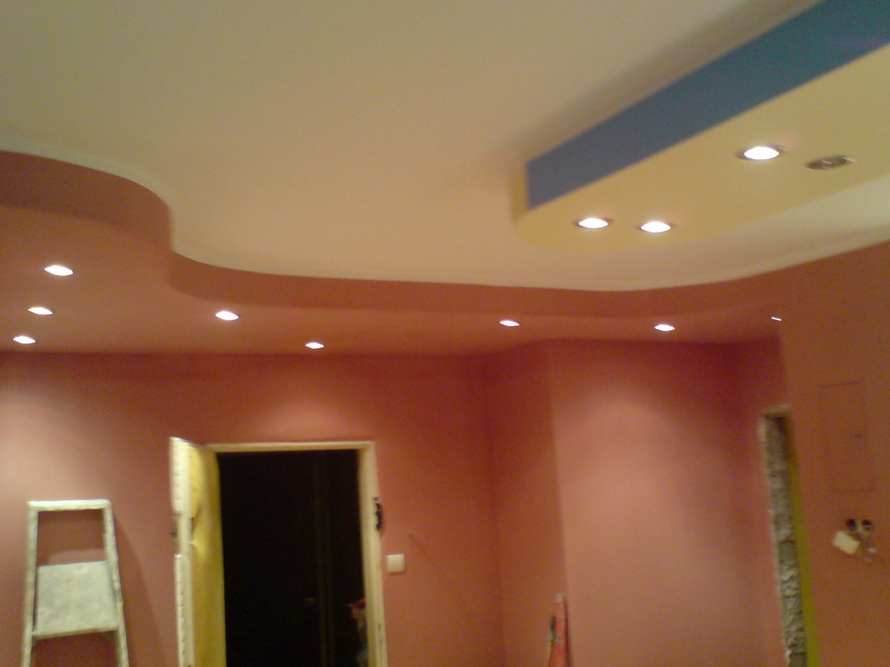 Wnętrze pokoju po remoncie z pomalowanymi na różowo ścianami, podwieszanym sufitem z oświetleniem punktowym i otwartymi drzwiami, widoczna drabina malarska.