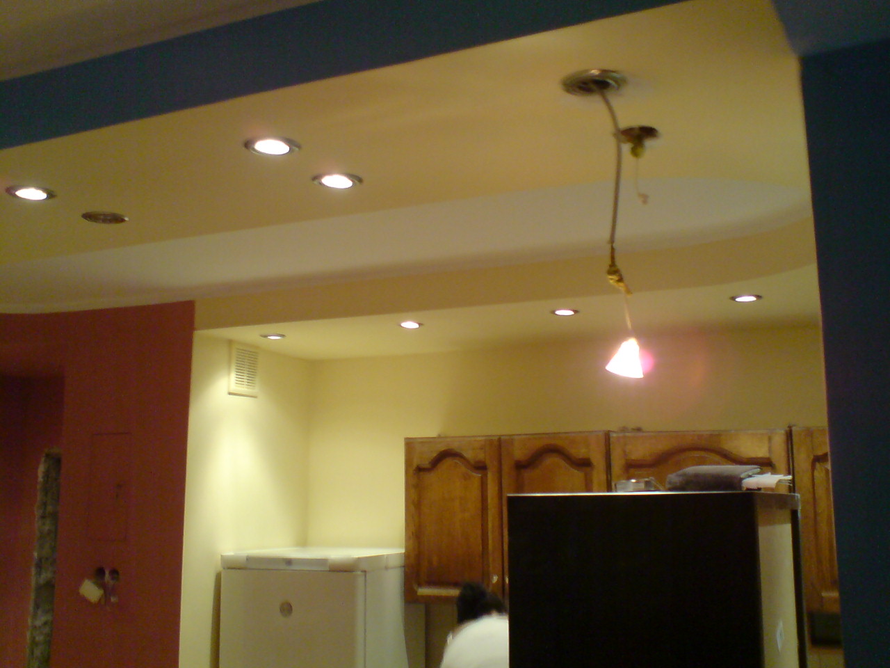 Wnętrze w trakcie remontu: widoczna instalacja elektryczna, sufit z oświetleniem punktowym, fragment ściany w kolorze ceglastym z widocznymi śladami prac, zabudowa kuchenna i sprzęt AGD.