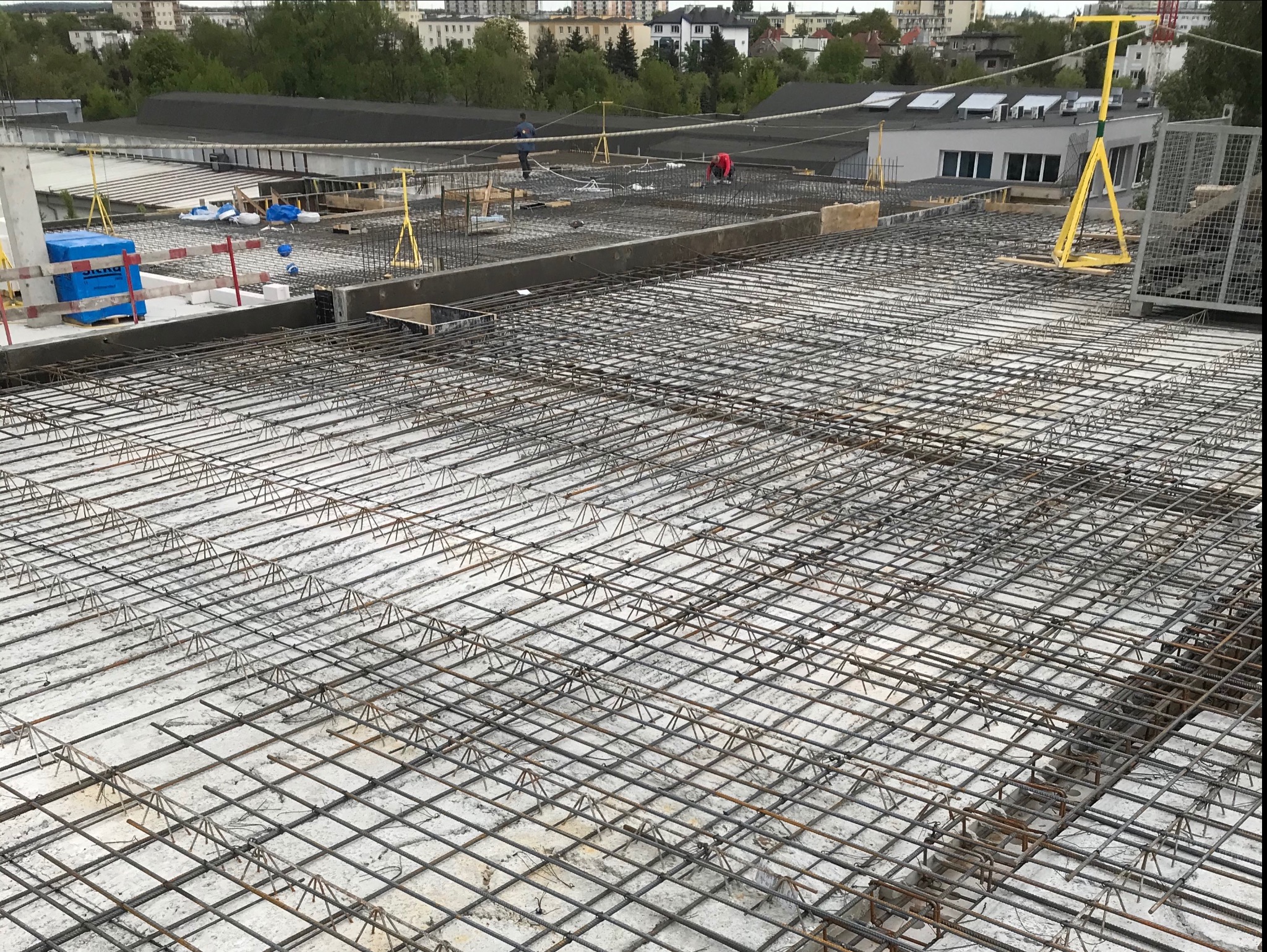 Siatka zbrojeniowa na betonowej płycie fundamentowej, z widocznymi dystansami i elementami szalunku na obrzeżach, w tle budynki i drzewa.