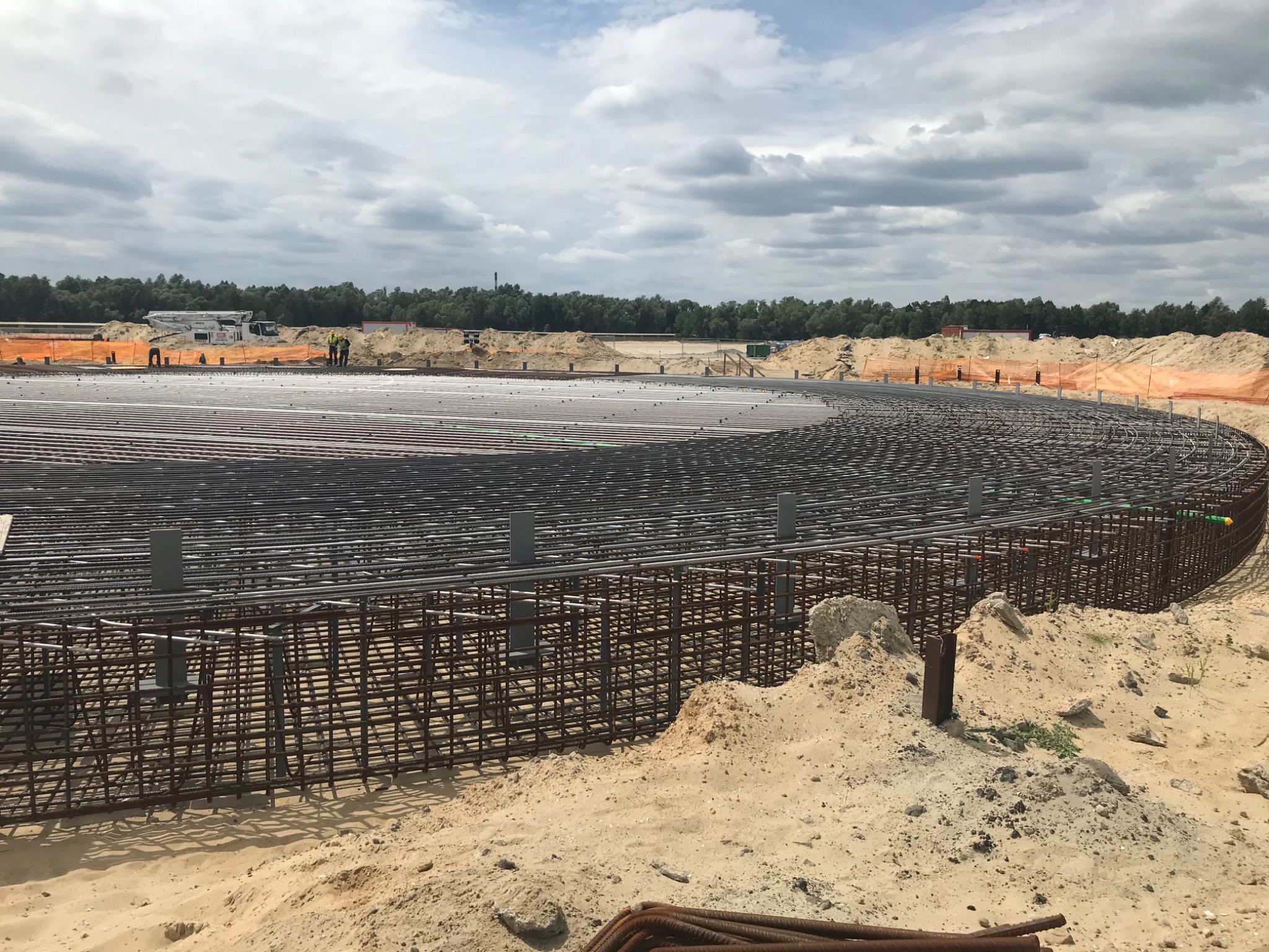 Rozległa, okrągła konstrukcja ze zbrojenia stalowego na piasku, przygotowana do zalania betonem; w tle widoczni pracownicy i sprzęt budowlany pod pochmurnym niebem.