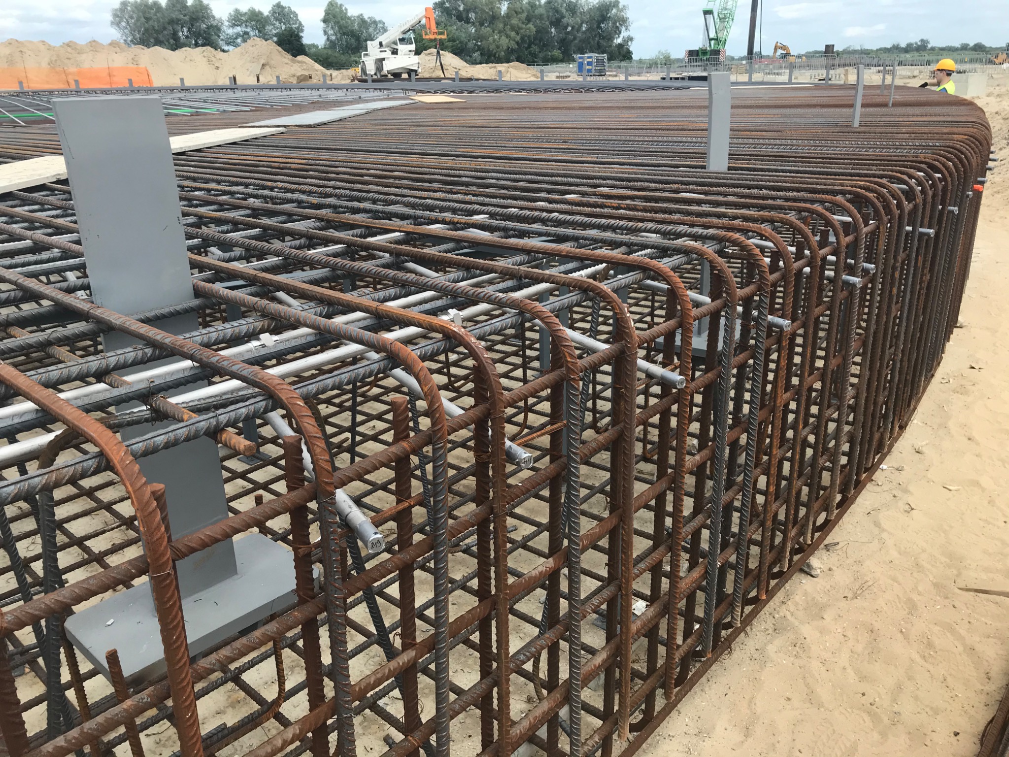 Siatka zbrojeniowa ułożona w zakrzywiony kształt na piasku, z szarymi wspornikami, widoczne elementy budowy w tle.