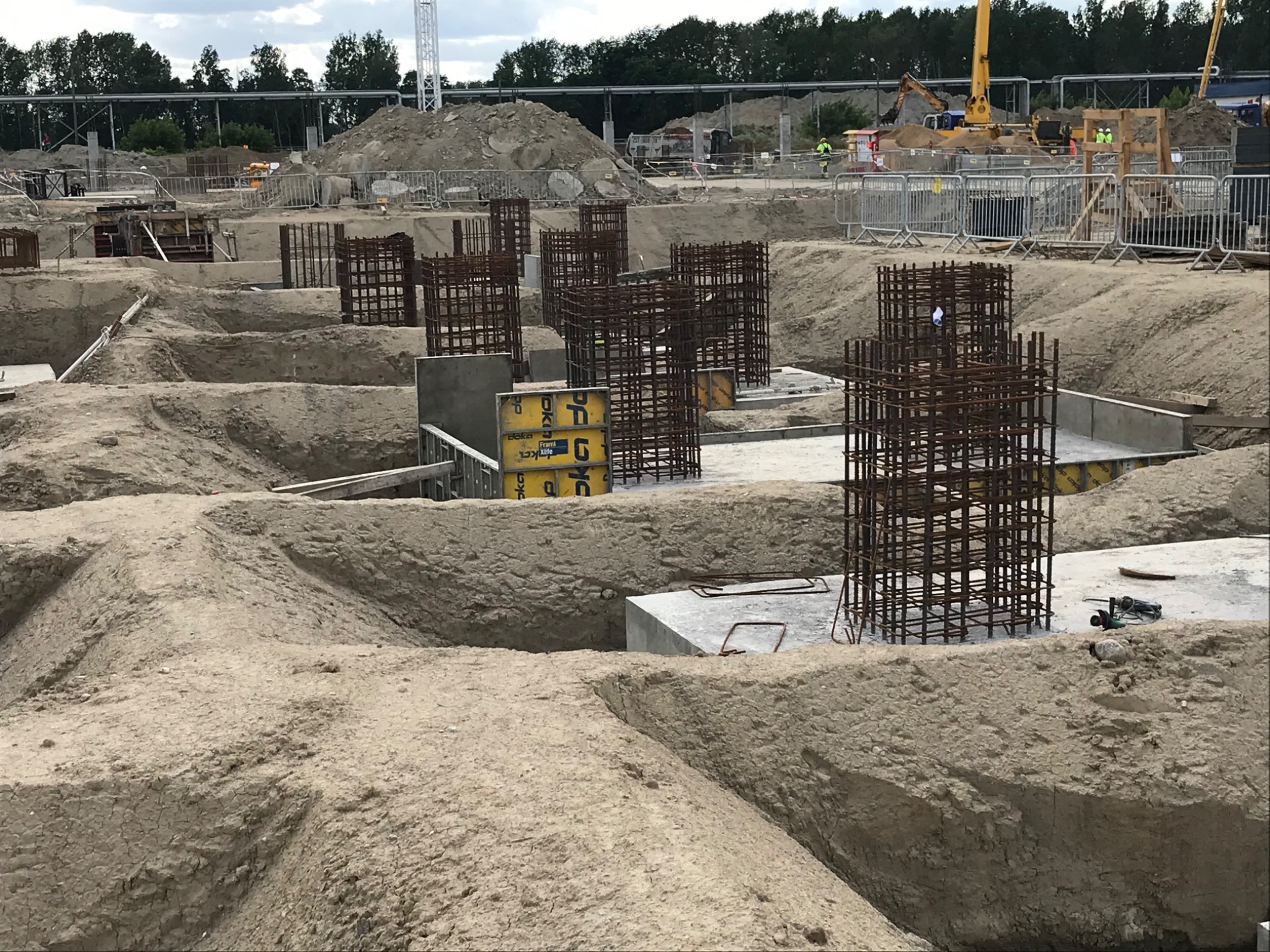 Przygotowanie fundamentów pod budynek: wykopy w ziemi, betonowe wylewki i wystające zbrojenia, na dalszym planie plac budowy z koparką i składowiskiem ziemi.