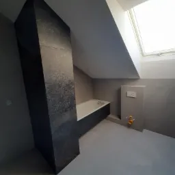 Łazienka na poddaszu w trakcie prac wykończeniowych: obudowana wanna, fragment pionowej ściany wyłożonej ciemnymi płytkami imitującymi metal, widoczne przyłącza kanalizacyjne i okno dachowe.