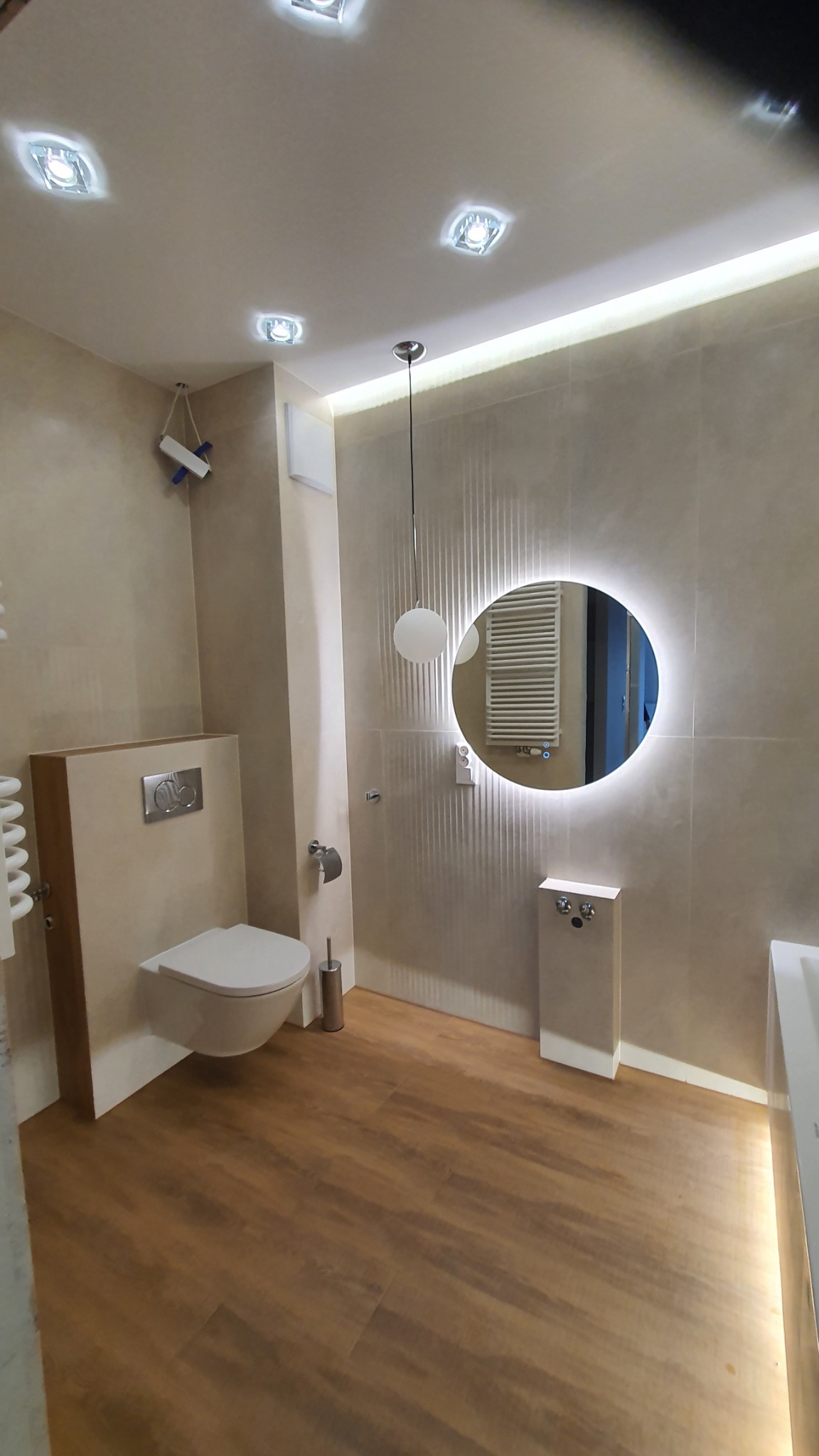 Nowoczesna łazienka z podświetlanym lustrem, drewnopodobną podłogą z paneli, beżowymi płytkami na ścianach i wiszącą toaletą ze spłuczką podtynkową.