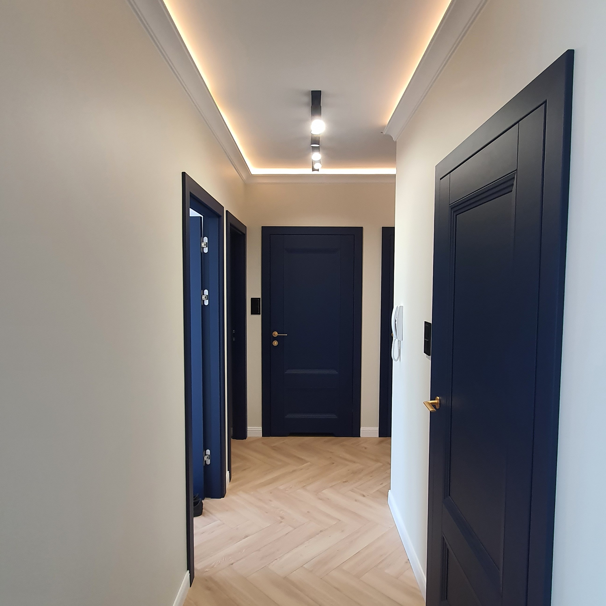 Korytarz po remoncie z trzema ciemnoniebieskimi drzwiami, parkietem ułożonym w jodełkę, listwami przypodłogowymi i oświetleniem LED podsufitowym.