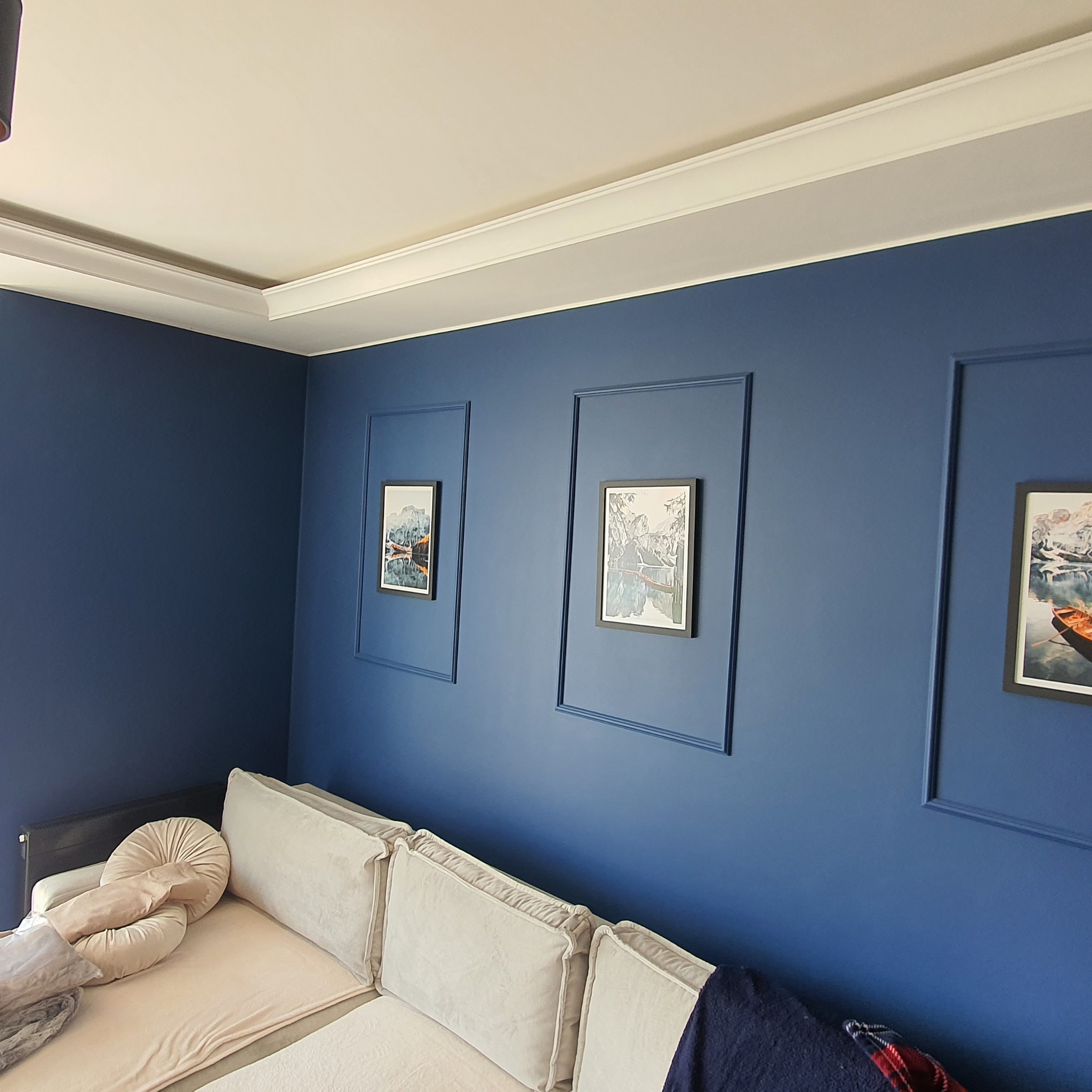 Granatowa ściana z dekoracyjnymi listwami tworzącymi ramy dla obrazów, kremowy narożnik z poduszkami i kocem w tle, biały sufit z ozdobną listwą.