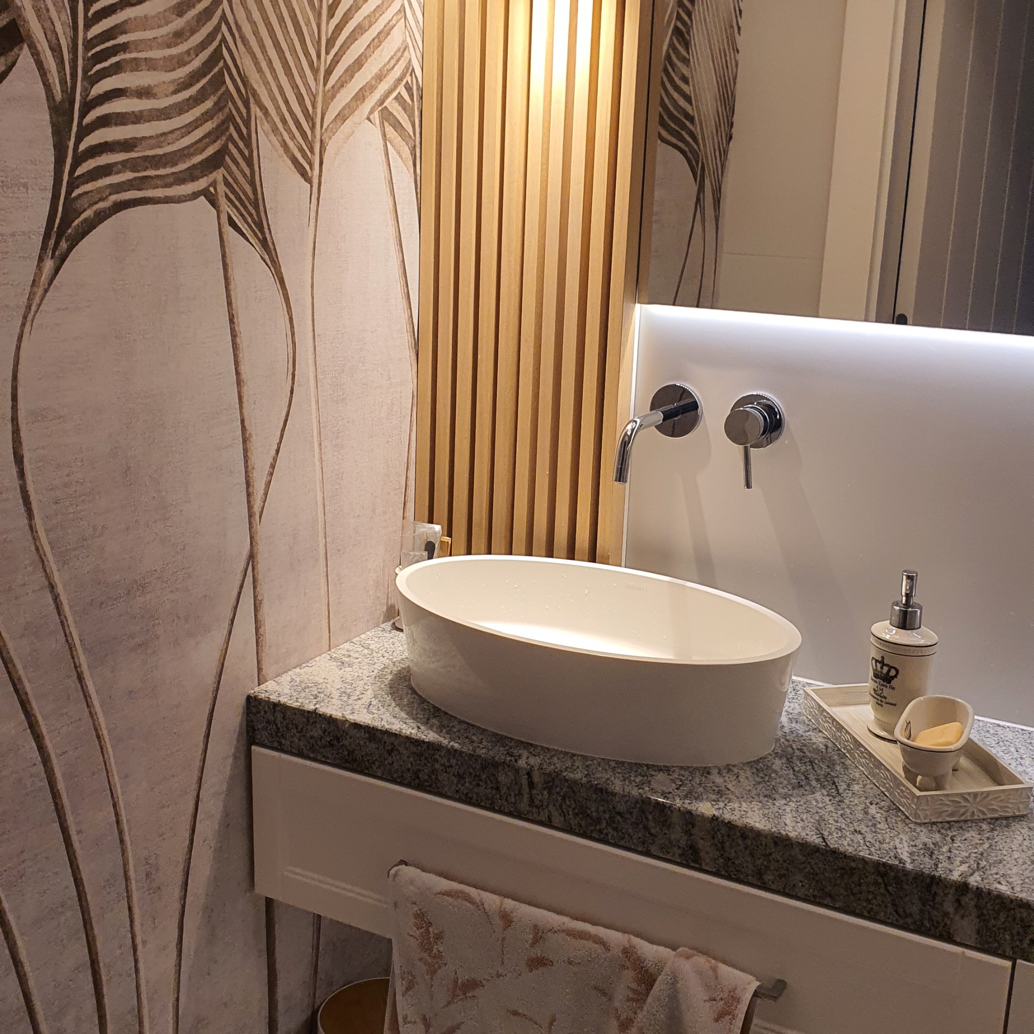 Nowoczesna łazienka z owalną umywalką nablatową na granitowym blacie, drewnianymi lamelami na ścianie i tapetą w motywy roślinne.