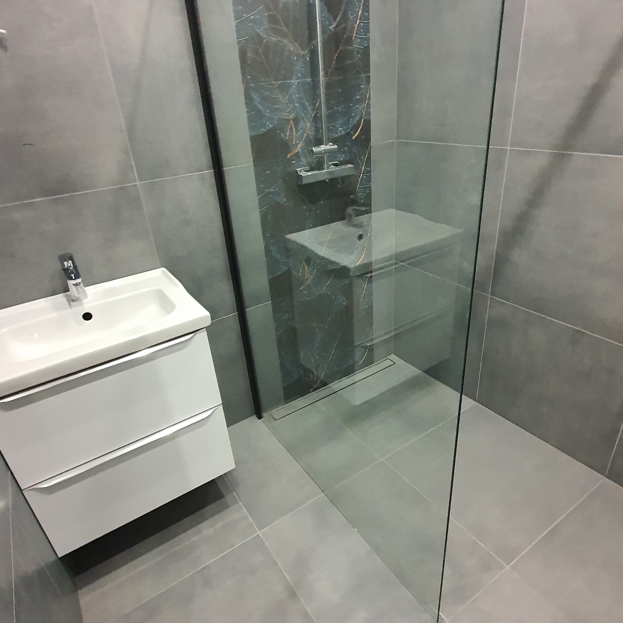 Nowoczesna łazienka z szarymi płytkami, kabiną prysznicową ze szklaną ścianką i umywalką z szafką w kolorze białym. Na ścianie pod prysznicem płytki z motywem liści.