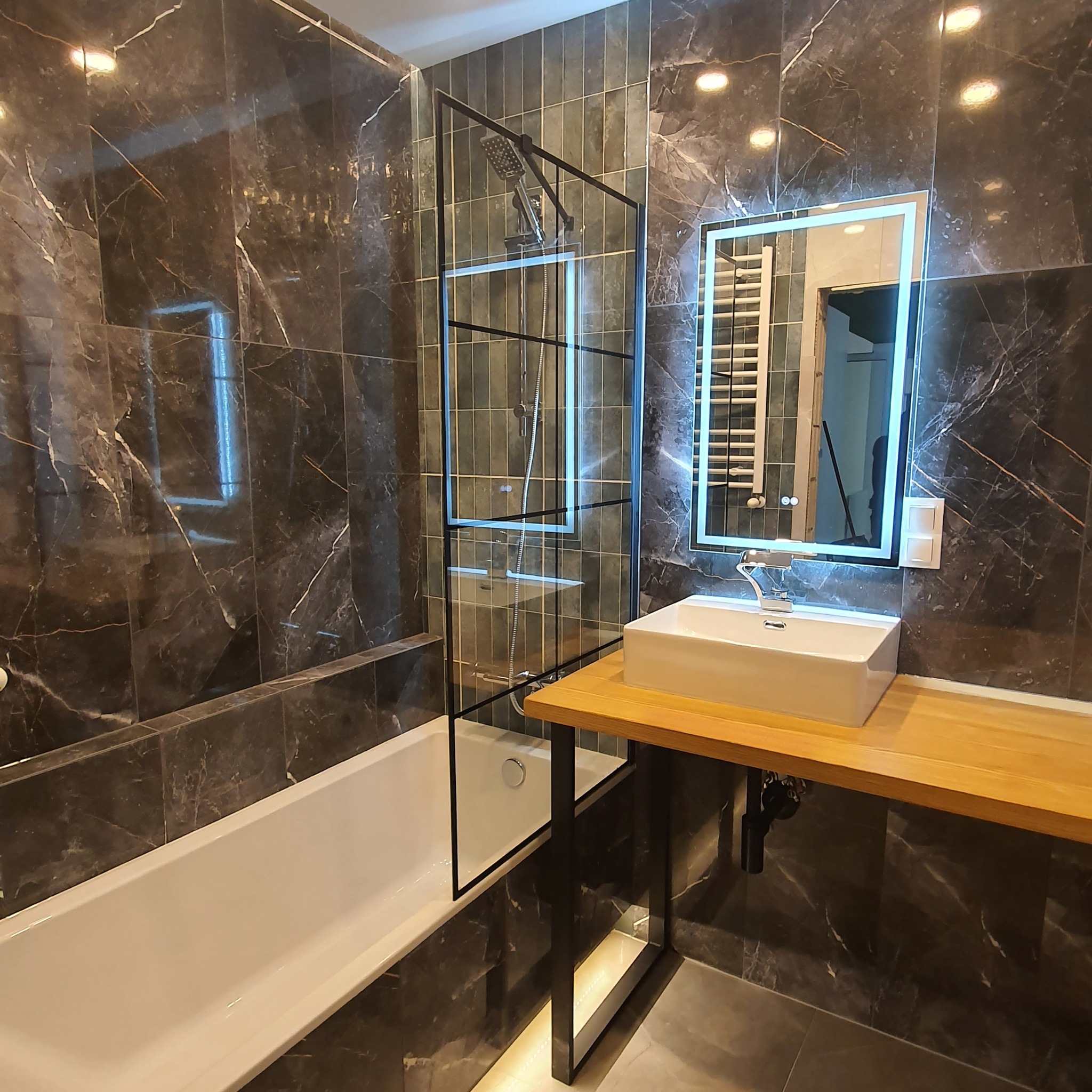 Nowoczesna łazienka z ciemnymi marmurowymi płytkami, białą wanną i umywalką nablatową na drewnianym blacie, oświetlonym lustrem LED i kabiną prysznicową ze szklaną ścianką.