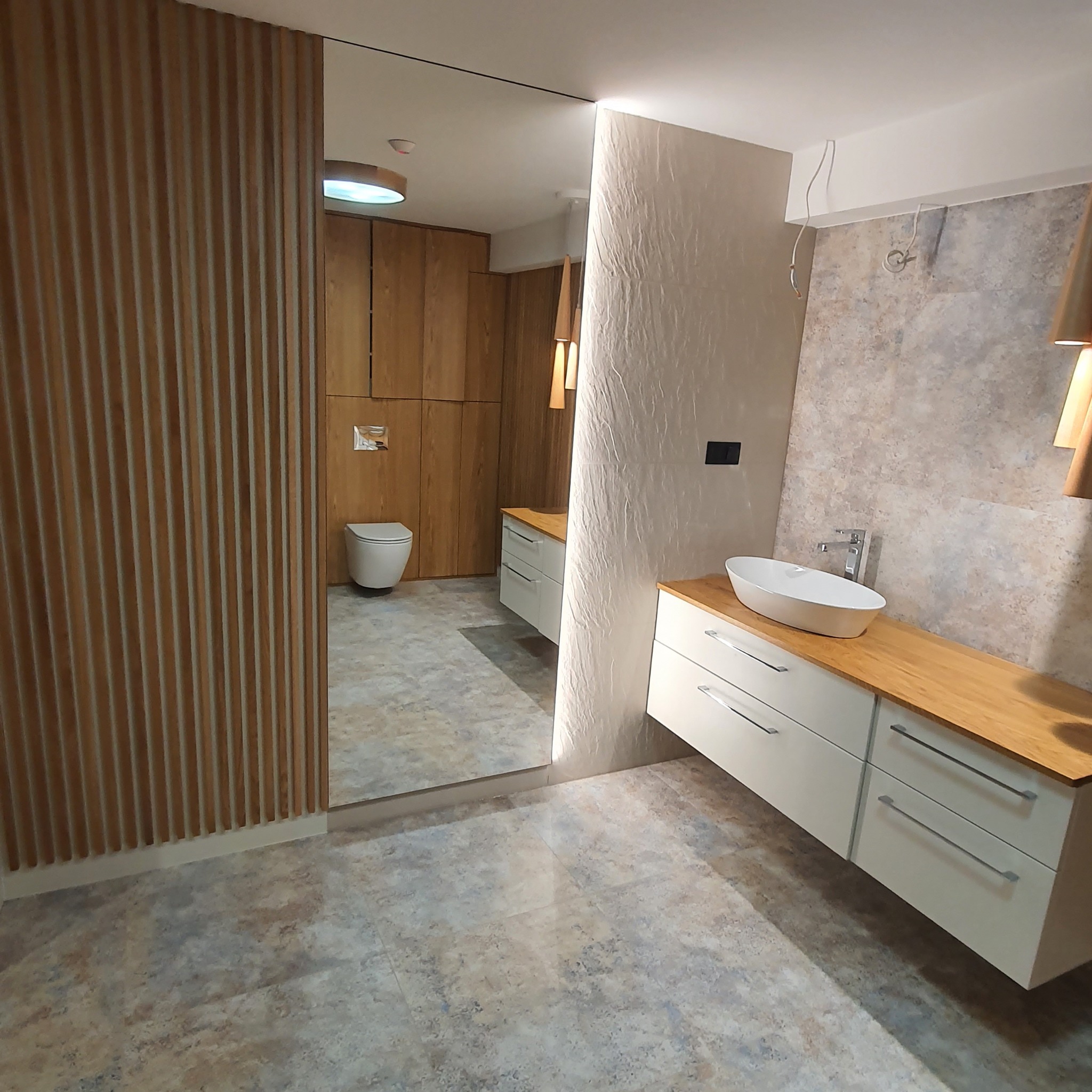 Nowoczesna łazienka z drewnianą zabudową, umywalką nablatową na blacie z jasnego drewna i płytkami imitującymi kamień. Lustro wkomponowane w drewniane lamele. Widoczne wiszące oświetlenie i toaleta...