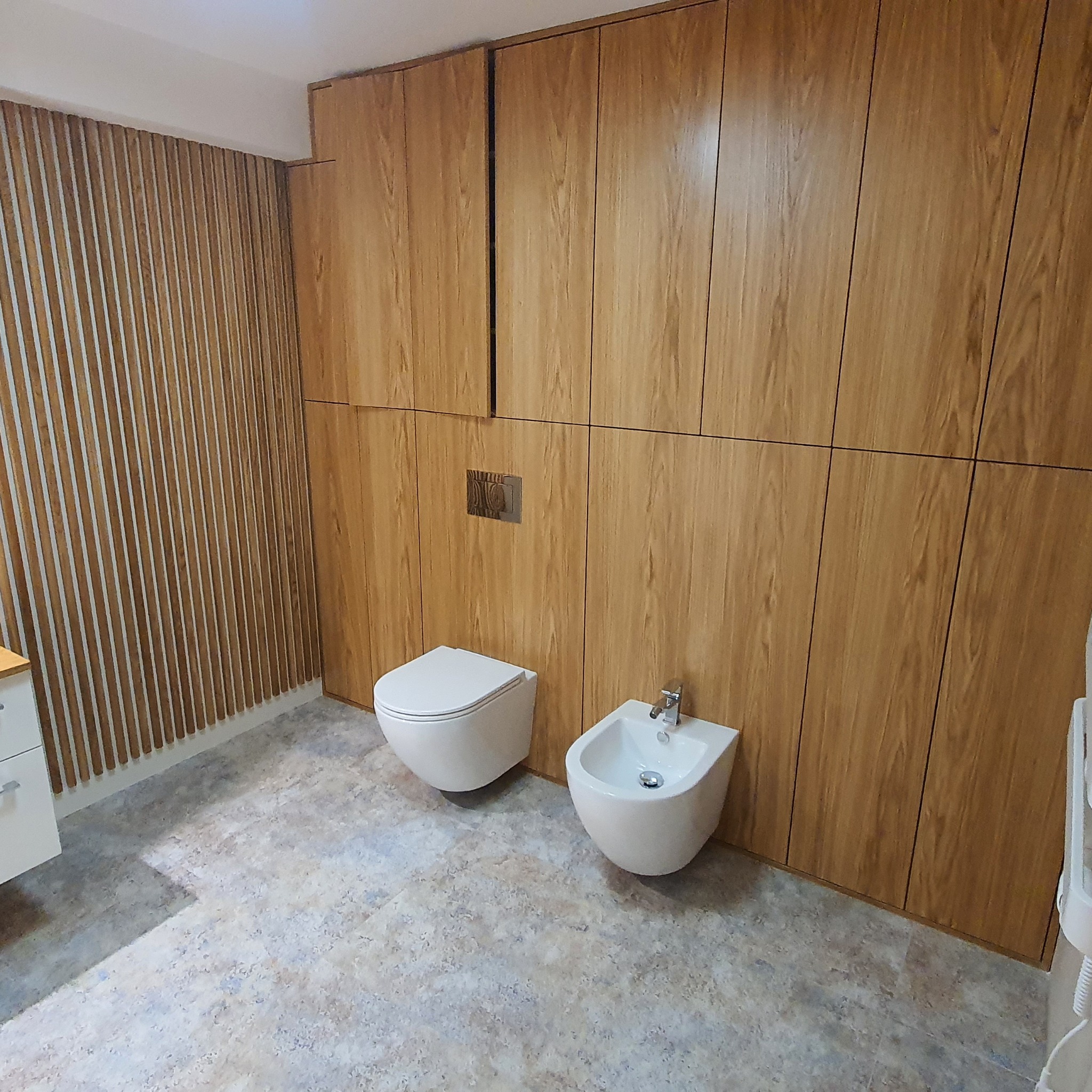 Nowoczesna łazienka z toaletą i bidetem na tle ściany z drewnianych paneli i szafek w zabudowie, szara podłoga imitująca kamień.