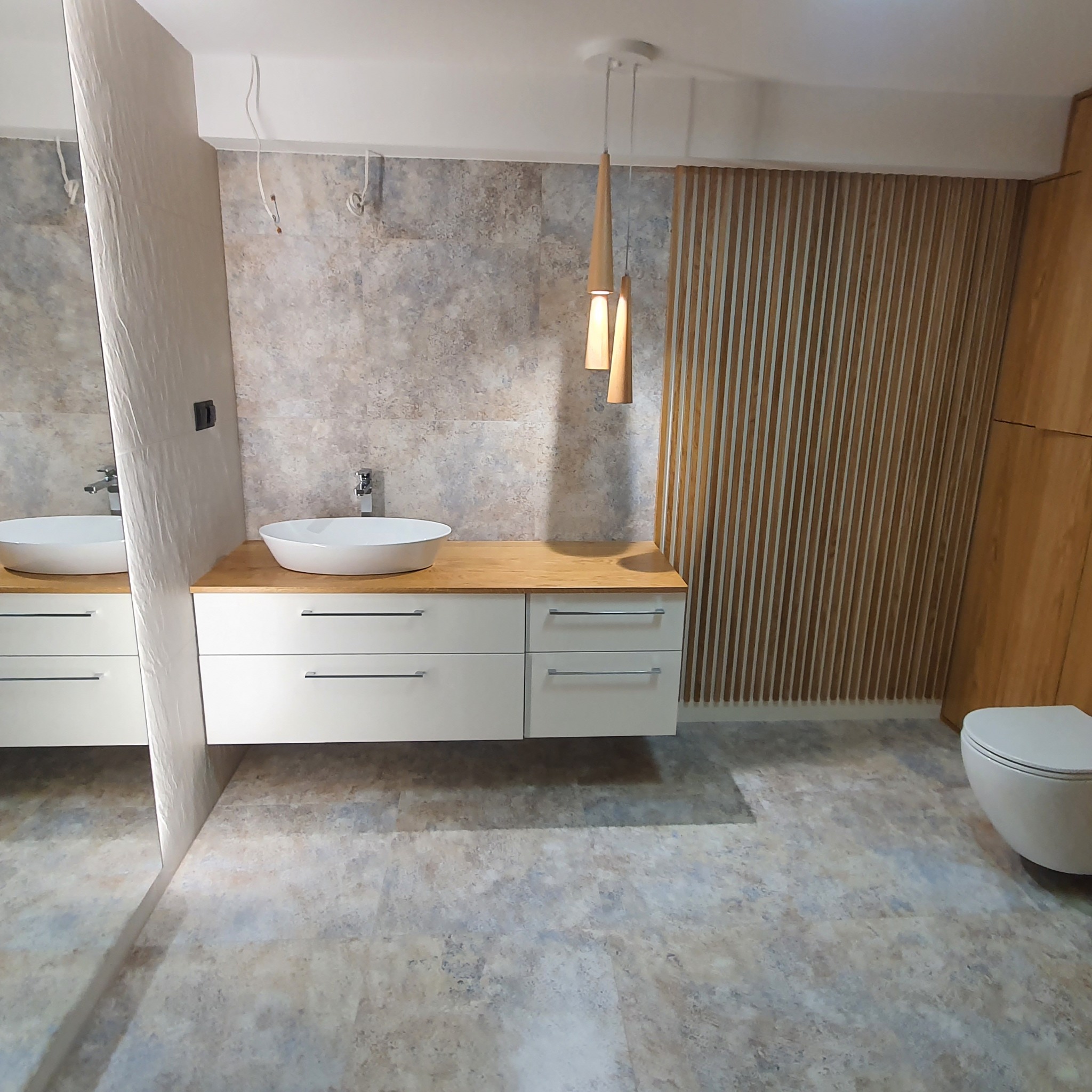 Nowoczesna łazienka z umywalką nablatową, drewnianym blatem, drewnianą ścianką działową z listewek, podwieszaną toaletą i lampami wiszącymi w kształcie stożków.