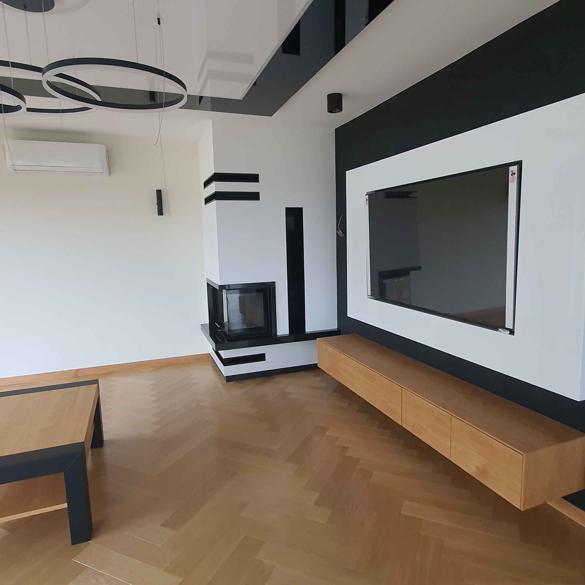Nowoczesny salon z parkietem w jodełkę, białymi ścianami, czarnymi akcentami, kominkiem z czarnymi paskami, podwieszanym telewizorem i drewnianą szafką RTV.