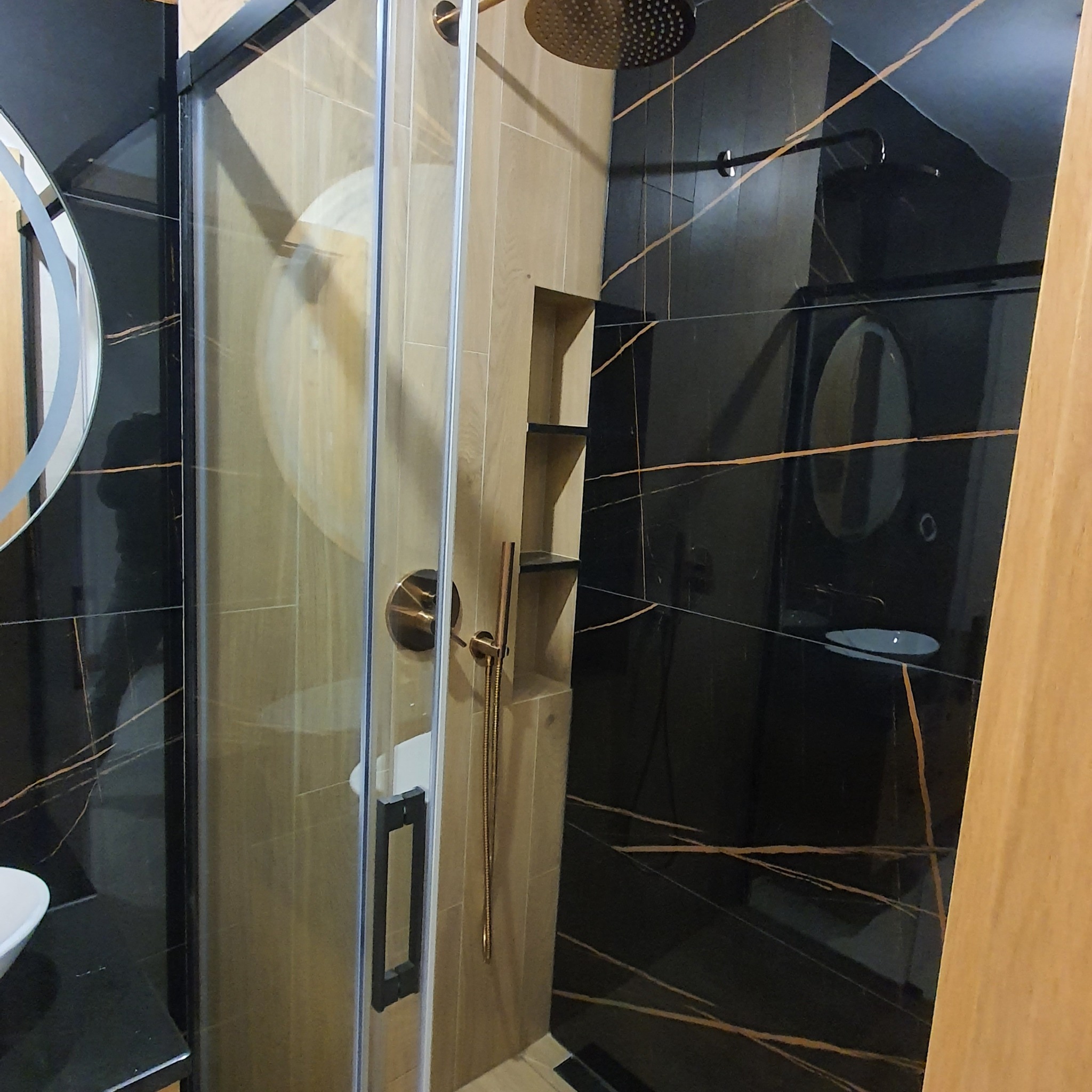 Nowoczesna kabina prysznicowa z deszczownicą, wykończona czarnymi płytkami z miedzianymi żyłkami i drewnopodobnymi panelami ściennymi z wnękami na kosmetyki.