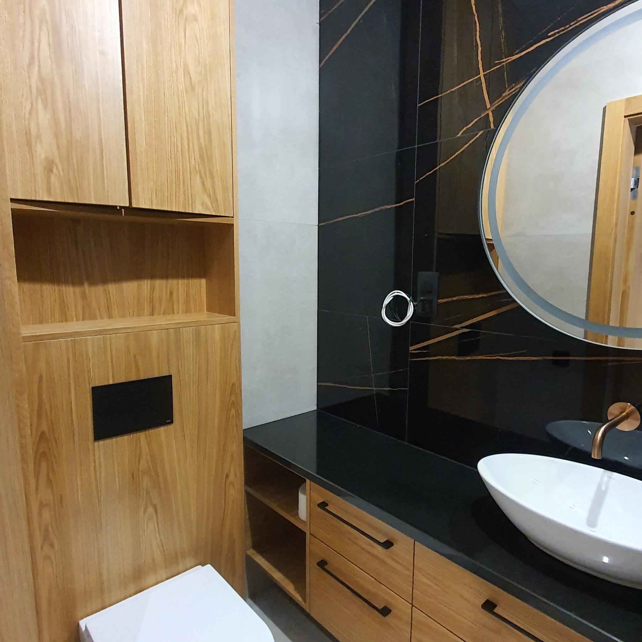Nowoczesna łazienka z drewnianymi szafkami, czarnym blatem i okrągłym lustrem z podświetleniem, akcentowana złotymi żyłkami w czarnej płytce ściennej.