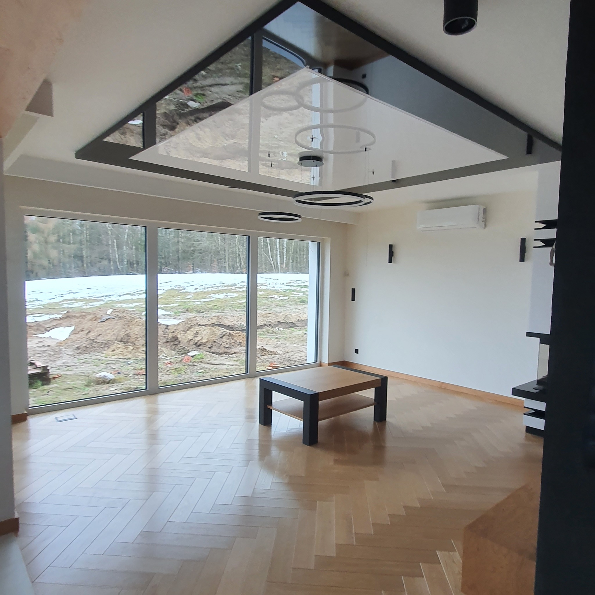 Jasny salon z parkietem ułożonym w jodełkę, dużymi oknami z widokiem na ogród w zimowej scenerii i nowoczesnym oświetleniem sufitowym z okrągłymi lampami.