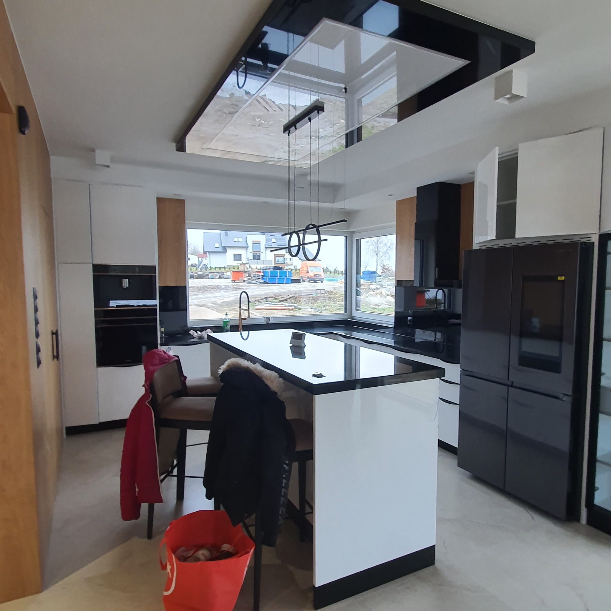 Nowoczesna kuchnia z wyspą, czarno-białe szafki, czarny blat, wisząca lampa z okręgami, widok na budowę za oknem.