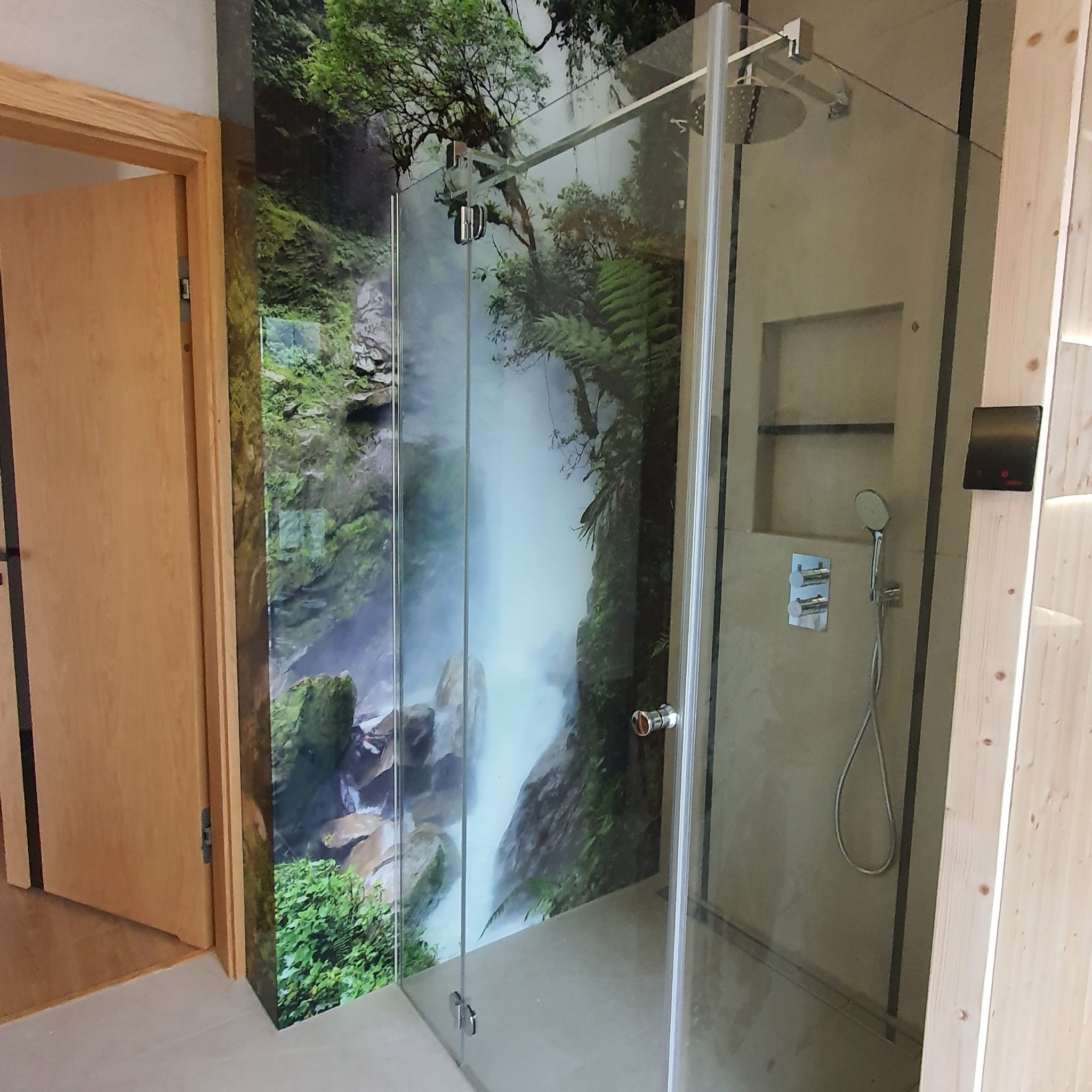 Nowoczesna kabina prysznicowa ze szklanymi ścianami, zamontowana na tle ściany z fototapetą przedstawiającą wodospad w lesie tropikalnym, widoczna armatura i panel sterowania.