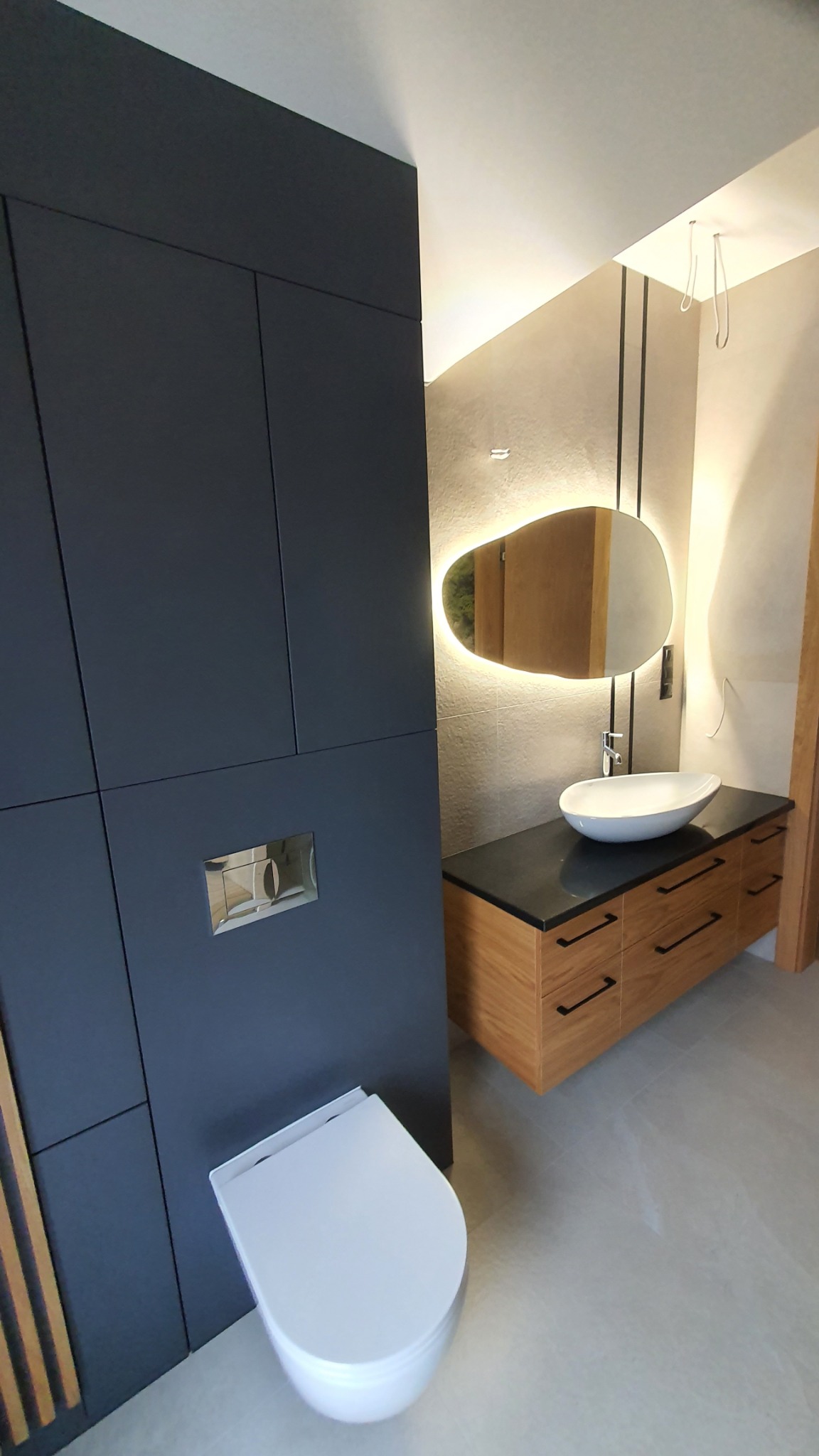 Nowoczesna łazienka z podwieszaną toaletą, szafką z umywalką nablatową i podświetlanym lustrem o nieregularnym kształcie.