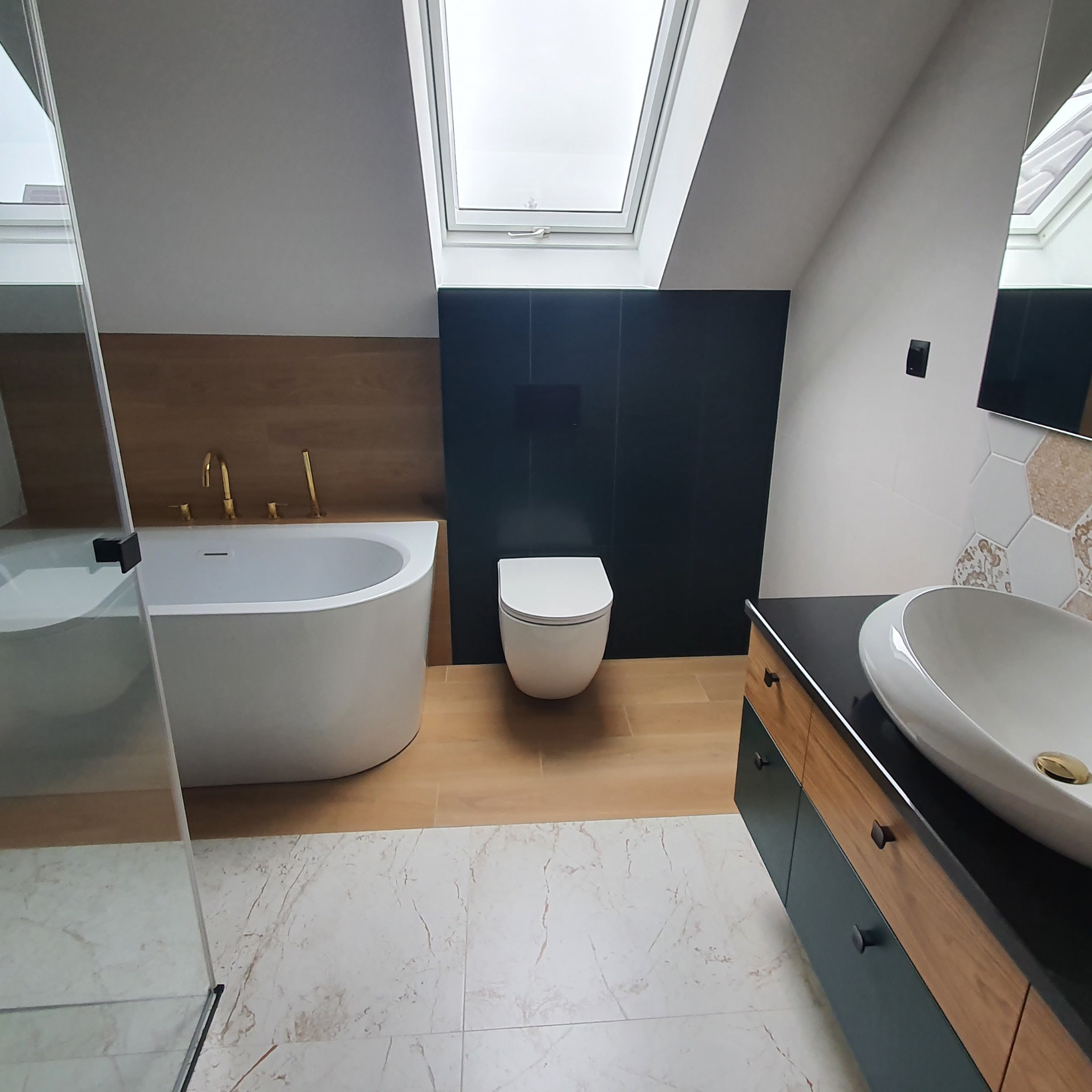 Nowoczesna łazienka na poddaszu z oknem dachowym, kabiną prysznicową ze szkła, wolnostojącą wanną z złotą armaturą, podwieszaną toaletą na tle ciemnych płytek oraz szafką umywalkową z blatem...