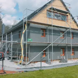 Dom w trakcie ocieplania elewacji styropianem grafitowym, z widocznymi rusztowaniami, żółtymi rurami wentylacyjnymi i niezabezpieczonym poddaszem, na tle trawnika i drzew.