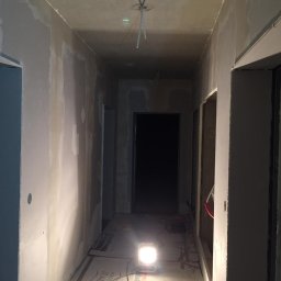 Bud-Men - Korytarz w trakcie remontu, ściany przygotowane do wykończenia, widoczne ślady po szpachlowaniu, na podłodze kartony ochronne i lampa robocza.