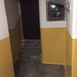 Bud-Men - Klatka schodowa z pomalowanymi na żółto ścianami do połowy wysokości, ciemne schody i drzwi na podeście, rura kanalizacyjna przy oknie.