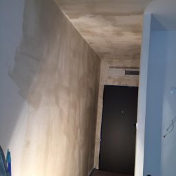 Bud-Men - Korytarz w trakcie remontu: ściany i sufit pokryte świeżo nałożoną warstwą szpachli, widoczna skrzynka elektryczna nad drzwiami, zabezpieczone niebieską taśmą malarską.