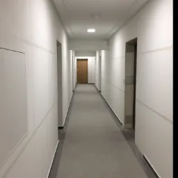 Długi korytarz w budynku biurowym lub mieszkalnym, świeżo po remoncie, z równo pomalowanymi ścianami na biało z szarymi pasami, szarą podłogą z płytek i drewnianymi drzwiami na końcu.