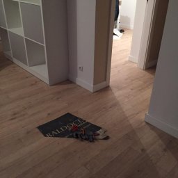 Bud-Men - Jasne panele podłogowe w korytarzu z widoczną okładziną ochronną i rękawicami roboczymi, częściowo widoczny regał i otwarte drzwi do innego pomieszczenia.