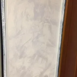 Ściana z efektem dekoracyjnym Jeger, wykończona w odcieniach szarości i bieli, z widoczną strukturą i delikatnym połyskiem, przylegająca do ramy okiennej i drewnianej futryny.