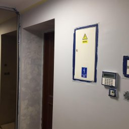 Bud-Men - Świeżo pomalowana ściana w odcieniach szarości z zabezpieczonymi taśmą malarską elementami elektrycznymi, widoczna faktura dekoracyjna obok futryny drzwi.