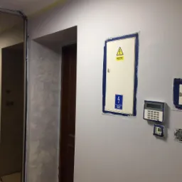 Świeżo pomalowana ściana w odcieniach szarości z zabezpieczonymi taśmą malarską elementami elektrycznymi, widoczna faktura dekoracyjna obok futryny drzwi.