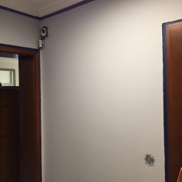 Bud-Men - Świeżo pomalowana na szaro ściana we Wrocławiu, zabezpieczona niebieską taśmą malarską przy suficie, ościeżnicach i grzejniku, z widocznym gniazdkiem elektrycznym bez obudowy.