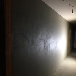 Bud-Men - Klatka schodowa w trakcie prac: ściana pokryta szarą warstwą wykończeniową, widoczne niedoskonałości i nierówności podłoża, surowe oświetlenie korytarza.