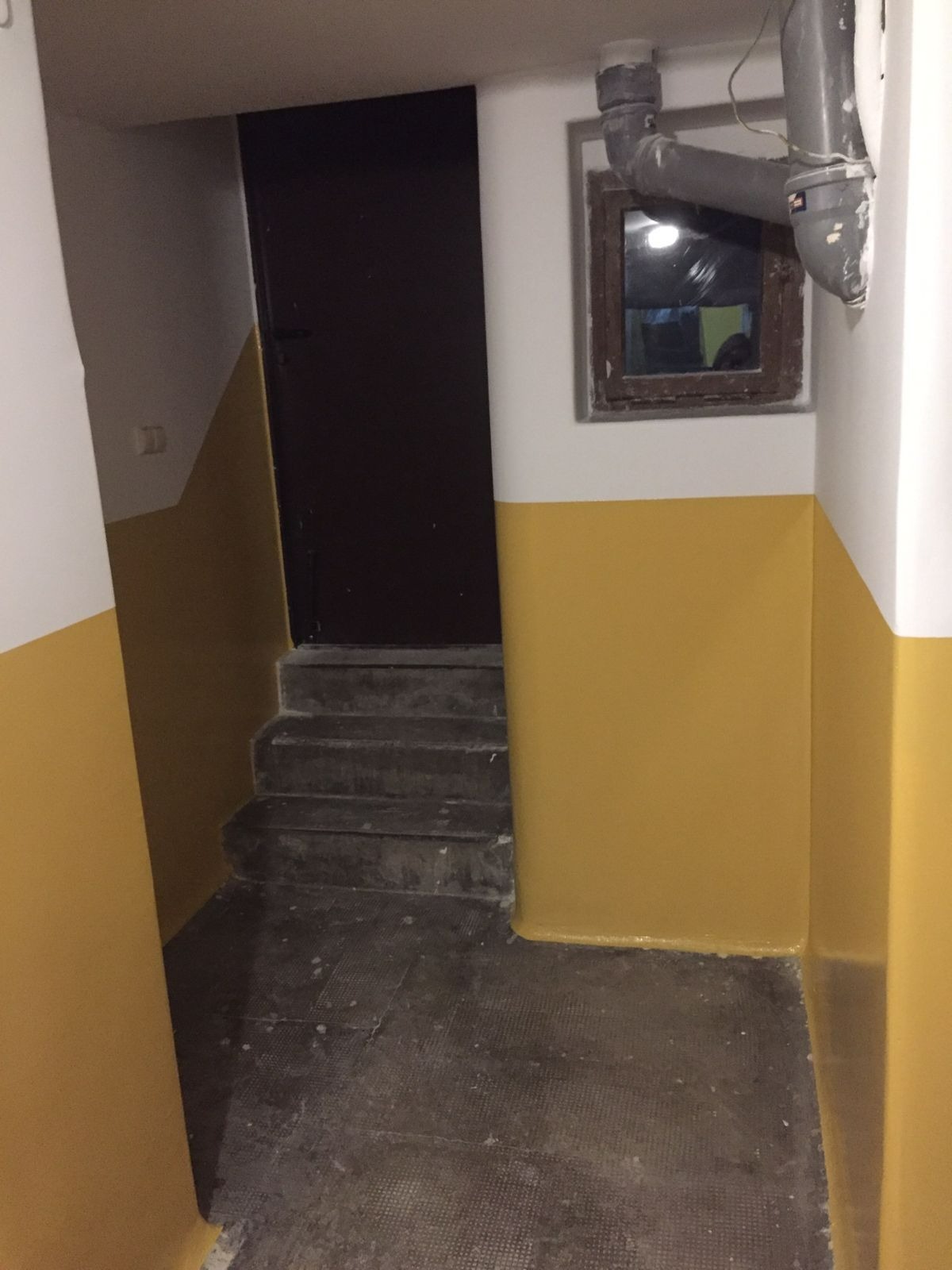 Klatka schodowa z pomalowanymi na żółto ścianami do połowy wysokości, ciemne schody i drzwi na podeście, rura kanalizacyjna przy oknie.