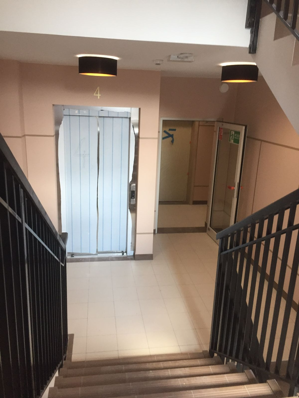 Klatka schodowa w bloku z widocznym wejściem do windy zabezpieczonym folią malarską oraz drzwiami ewakuacyjnymi, oświetlona dwoma okrągłymi lampami sufitowymi.
