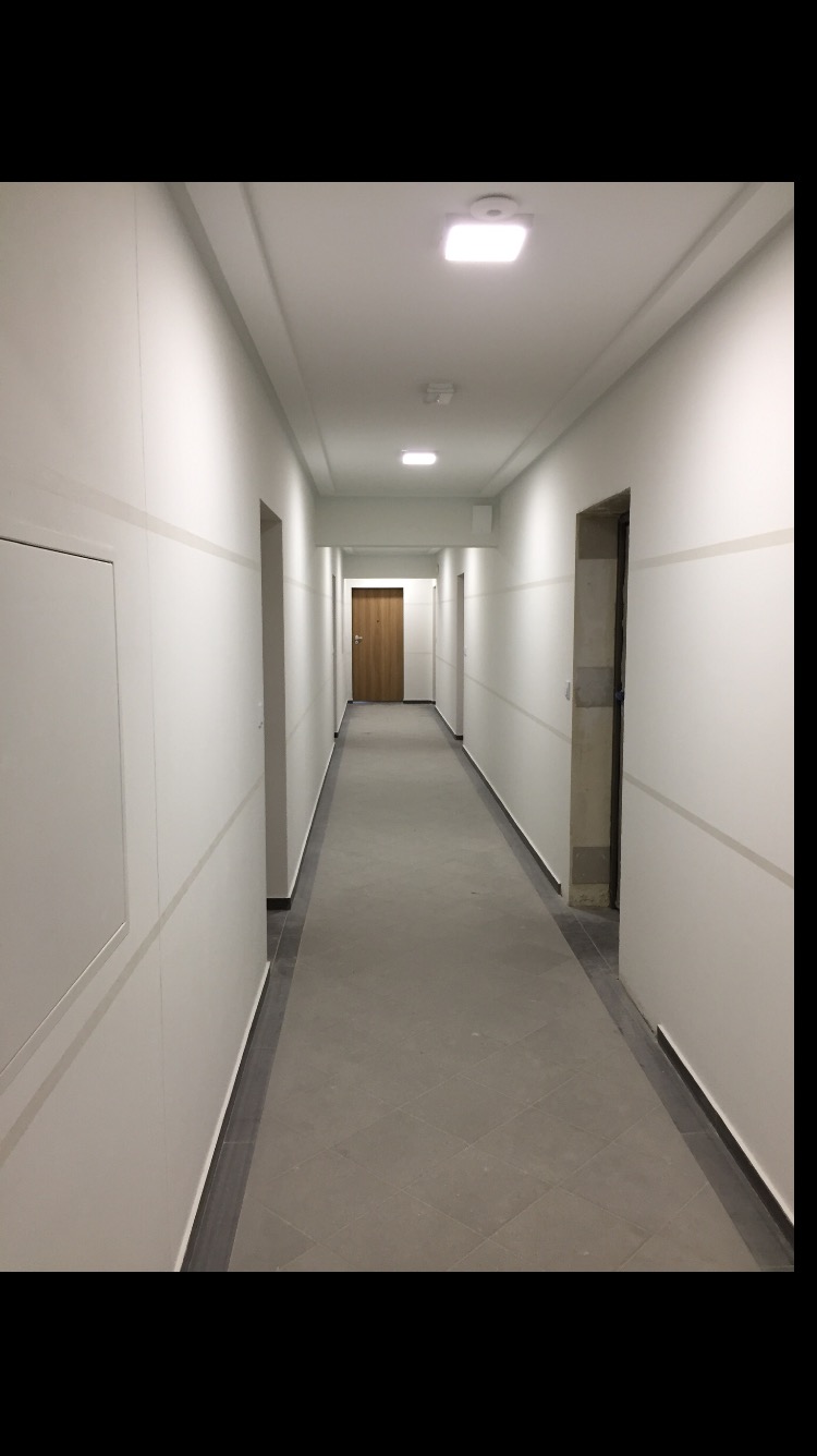 Długi korytarz w budynku biurowym lub mieszkalnym, świeżo po remoncie, z równo pomalowanymi ścianami na biało z szarymi pasami, szarą podłogą z płytek i drewnianymi drzwiami na końcu.